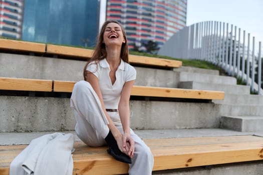Joyful young woman in casual white clothing laughing and sitting on wooden steps in an urban outdoor setting with modern city skyline in background, expressing happiness and freedom.