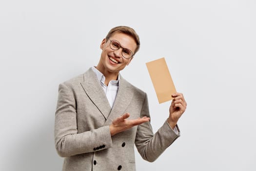 Smiling man in glasses and beige blazer holding paper card, gesturing with hand, isolated on white background. Casual business style, cheerful expression, modern communication and remote digital freelancer concept.