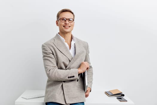 smiling young caucasian man wearing glasses and grey knitted jacket hold laptop while stand near table with smartphone and notebook on plain white background. technology freelance work and digital nomad lifestyle concept.