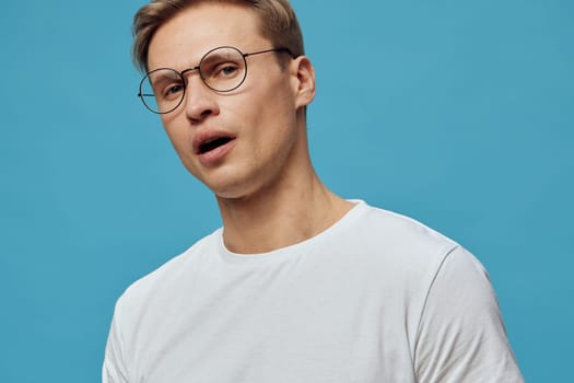 portrait of young caucasian man wearing round glasses and white t-shirt looking surprised or shocked against plain blue background. expression shows emotional reaction during digital detox or mental health break from gadgets. Concept: mental health and gadgets