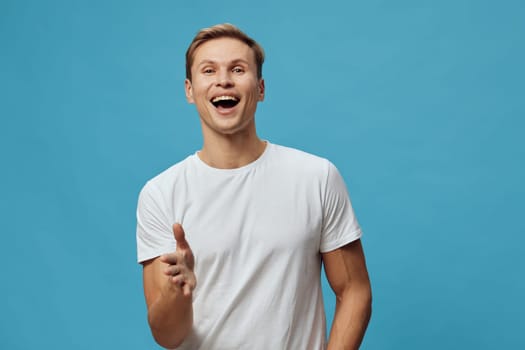 happy smiling man wearing casual white t-shirt standing against blue background showing cheerful joyful facial expression with open mouth and laughter in studio portrait. Concept: positive emotion and happiness