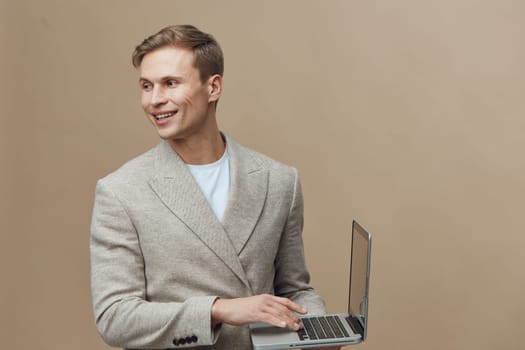 Smiling young caucasian man wearing beige jacket holding laptop looking aside on plain beige background. The man represents digital nomad lifestyle and remote work concept with casual modern style. Concept: digital nomad work
