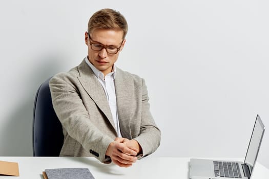 young man stretching hands while sitting at white desk with open laptop and notebook wearing casual blazer and glasses focused on work digital nomad lifestyle and remote work in metaverse concept
