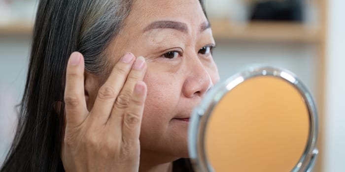 An elderly woman applies skincare cream to her face, focusing on her beauty and wellness as she enjoys her skincare routine in front of a mirror.