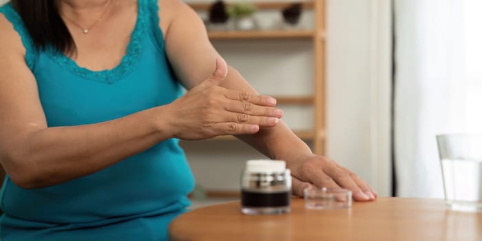 An elderly woman enjoys her beauty routine, applying cream to her arm in a serene home environment, emphasizing the importance of wellness and skincare.