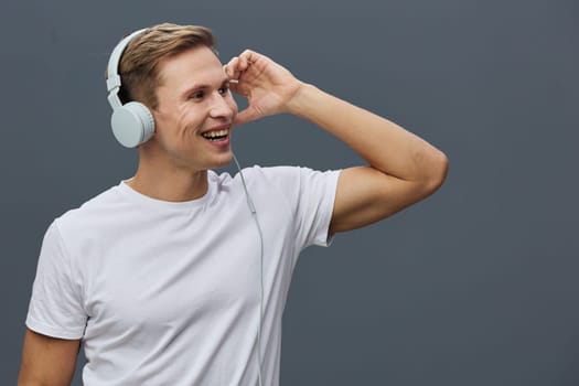 Smiling young man wearing white headphones and casual white t-shirt looking sideways while listening to music or participating in video calls. Modern gray background enhances focus on cheerful expression and relaxed pose. Concept: digital nomad lifestyle