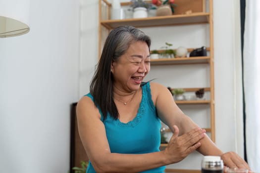 A happy elderly woman applies skincare cream to her arm, enjoying her beauty routine in a cozy home setting, highlighting the joy of self-care.