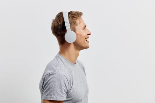 Smiling young caucasian man in grey t-shirt wearing white headphones looking happy and relaxed while enjoying music and mental health and gadgets concept isolated on plain light background studio portrait