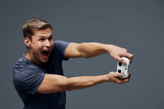 excited young man holding wireless game controller wearing casual t-shirt standing against dark gray background with energetic facial expression, digital detox and entertainment concept