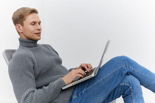 relaxed man using laptop in casual grey sweater and blue jeans sitting on modern chair focused on screen digital nomad working remotely with wireless internet lifestyle concept