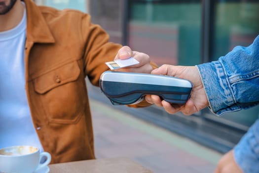 Close-up of two people making a contactless card payment at a cafe table with a POS terminal. Coffee and pastry are visible. Modern lifestyle and digital finance concept in urban setting.