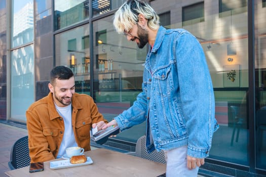 Caucasian Man pays with a contactless card at an outdoor cafe while his friend watches and smiles. Modern street cafe scene with relaxed atmosphere and casual fashion in an urban environment.