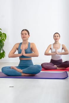 Caucasian women practicing yoga in seated lotus pose with palms together, wearing sportswear on mats in a bright indoor space.Calm expression and focus reflect mindfulness and healthy lifestyle.
