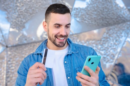 Happy young man using his smartphone while holding a credit card. He smiles with excitement, standing outside a glass building, enjoying an online shopping or payment experience.