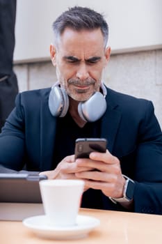 Mature businessman focused on smartphone at outdoor cafe table. Headphones around neck, coffee and tablet on table. Vertical format with modern professional style and urban background.
