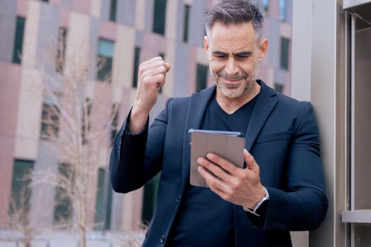 Confident businessman celebrates winning an online bet, holding a tablet and raising his fist. He smiles outdoors in a city setting. Concept of success, digital lifestyle, and modern finance.