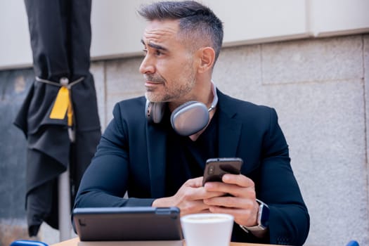 Mature businessman with headphones around neck sits at a cafe table holding a smartphone. Tablet and coffee on the table. He looks aside with a thoughtful expression in a modern urban setting.
