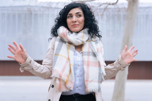 Portrait of a woman wearing a plaid scarf and beige coat, standing outdoors with raised hands. Confident expression in a cold urban setting. Concept of winter fashion, attitude, and individuality.