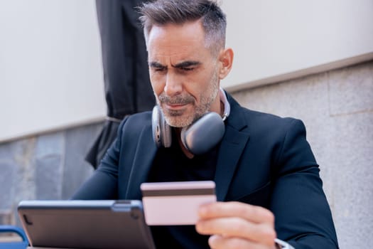 Confident caucasian man with headphones smiling while using tablet and holding a credit card outdoors. Concept of online shopping, digital payment, modern lifestyle, and financial technology.