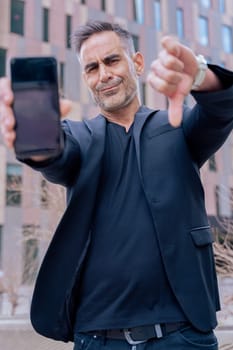 Businessman in black outfit shows his smartphone and gives a thumbs down He serious confidently in an urban background, suggesting success and desapproval of modern technology looking at camera