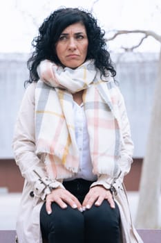 Horizontal portrait of a woman sitting on a bench outdoors. She wears a beige coat and scarf, looking calm and confident. Urban background with winter atmosphere and seasonal fashion concept. Vertical