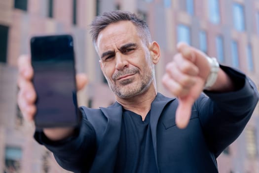 Businessman in black outfit shows his smartphone and gives a thumbs down He serious confidently in an urban background, suggesting success and desapproval of modern technology looking at camera