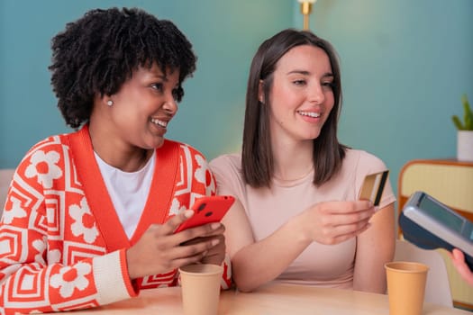 African American and Caucasian women smiling while paying at a coffee shop. One holds a card, the other a phone. Concept of friendship, technology, and casual lifestyle in a modern setting.