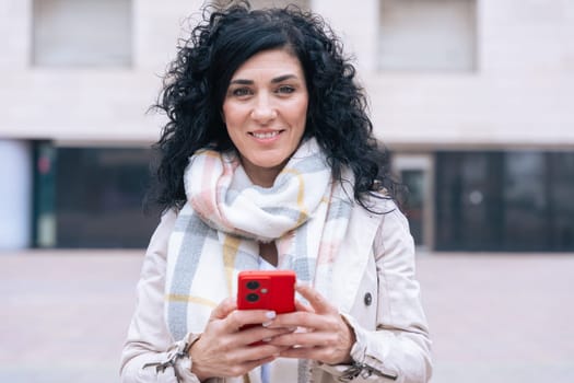 Happy caucasian woman standing outdoors in urban setting,wearing coat and scarf,using her red smartphone.She is smiling and relaxed,enjoying a casual moment on a cold day in the city looking at camera