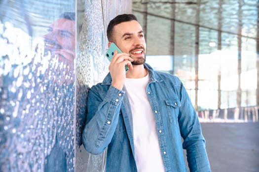 Smiling man in denim shirt talks on his smartphone while leaning against a glass wall. Bright daylight and architectural reflections create a clean, modern and relaxed business or lifestyle scene.