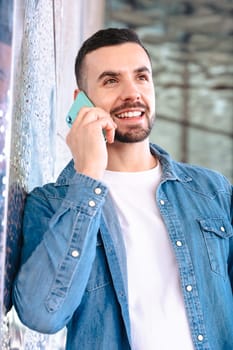 Smiling man in denim shirt talks on his smartphone while leaning against a glass wall. Bright daylight and architectural reflections create a clean, modern and relaxed business or lifestyle scene.