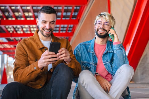 Two friends sit together outdoors, laughing and pointing at a smartphone screen. Casual outfits and red architectural background suggest a modern, social and relaxed moment in the city.