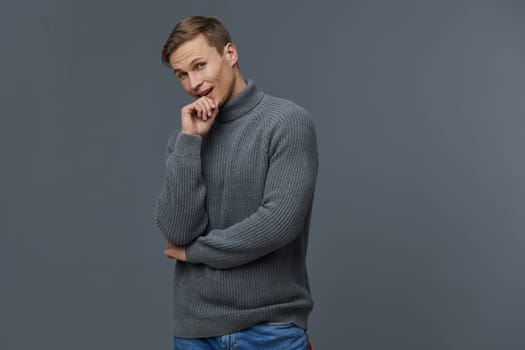 young caucasian man thoughtful in grey knitted sweater and blue jeans standing against grey background with hand on chin, contemplating automation tasks and digital work. Concept: automation tasks and remote work.
