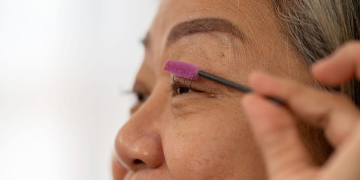 An elderly woman carefully brushes her eyebrows, highlighting her attention to detail in her beauty regimen and self-care.