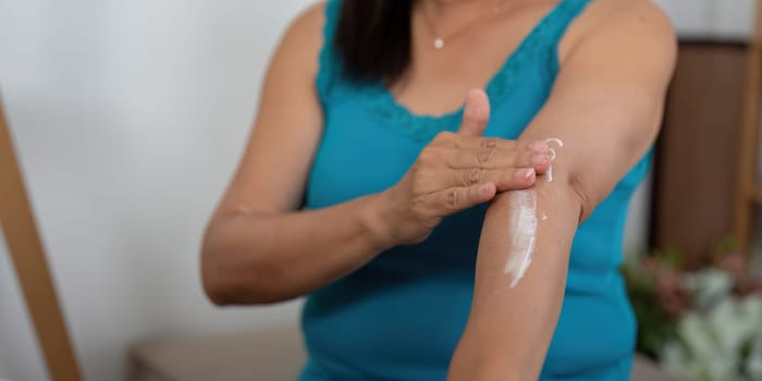 An elderly woman enjoys a moment of self-care, applying lotion to her arm while seated comfortably at home. This image highlights the importance of beauty and wellness routines at any age.