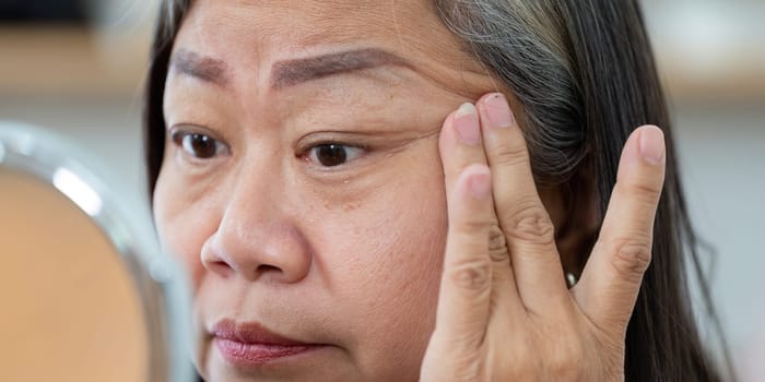 A senior woman is focused on applying cream around her eyes, highlighting her commitment to beauty and wellness through skincare.