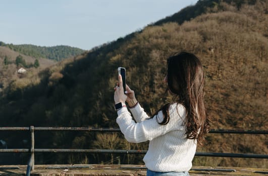 Portrait from the waist up sideways of a young beautiful brunette girl who is filming a mountain landscape with a mobile phone standing on a viewing platform in an ancient castle, a spring sunny day, a side view close-up. Concept using technology, time of travel and tourism.