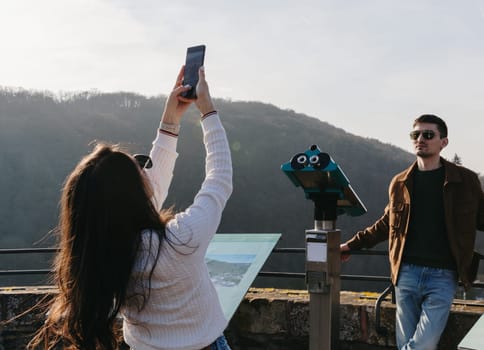 A waist-up portrait of a young girl from behind, half-turned, who is photographing a beautiful guy with a mobile phone from an observation deck in an ancient castle, a spring sunny day, a side view, close-up. Concept using technology, time of travel and tourism.