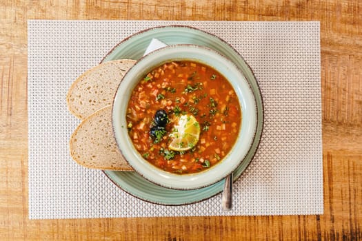 Hot homemade solyanka soup with olives, lemon wedge and chopped parsley in a modern beautiful plate stand on a wicker napkin on a wooden table in a restaurant, flat lay close-up. Concept traditional dishes.