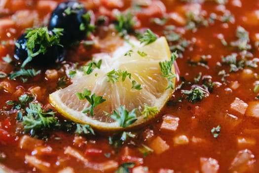 beautiful appetizing view of lemon wedge with olives in hot homemade solyanka soup with chopped parsley in modern beautiful plate stand on table in restaurant,top view from side hyper close-up.Concept traditional dishes.
