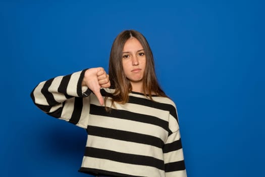 Young brunette Hispanic teenager wearing a striped sweater looking unhappy and angry, showing rejection and negativity with a thumbs-down gesture, isolated over blue background. Bad expression