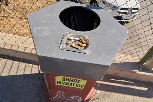 Crete.Greece - april 23, 2025: Street ashtray in Heraklion with a metal plate for cigarette disposal. A yellow municipal sign is visible, placed next to a chain-link fence with parked vehicles in the background.