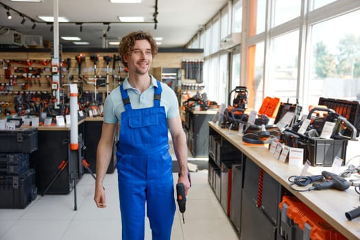 Workman wearing coveralls walking with new drill among hardware store showcase being satisfied with good purchase