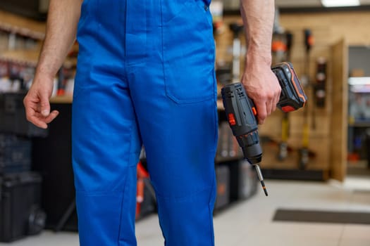 Crop portrait of repairman wearing coveralls holding drill power tool in hands standing over workshop interior. Craftsman providing high quality service of hardware repair and maintenance