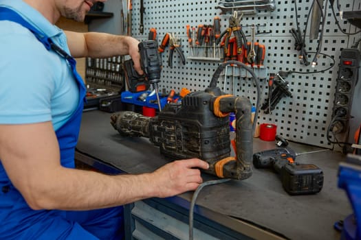 Handyman examining broken perforator power tool before starting to repair. Master providing inspection and inspection of damaged dirty puncher standing at worktable in workshop