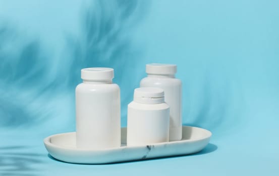 Three white plastic pill bottles of varying sizes stand on a white marble tray against a light blue background with subtle leaf shadows