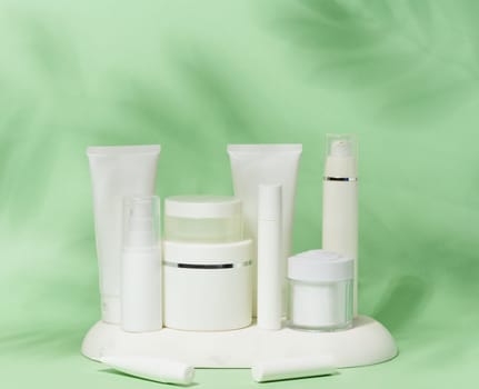 A collection of various white cosmetic containers, including tubes, jars, and bottles, are arranged on a white stand against a light green background with leaf shadows.
