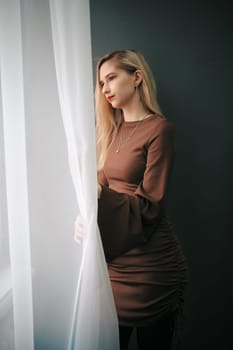 A young woman stands by a window with sheer curtains, looking out contemplatively.