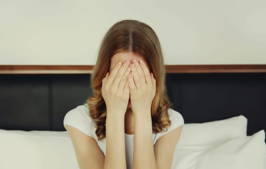 Upset sad crying young woman covering her eyes and face with her hands while experiencing depression, stress, fatigue, mental suffering after sleeping sitting on the bed at home