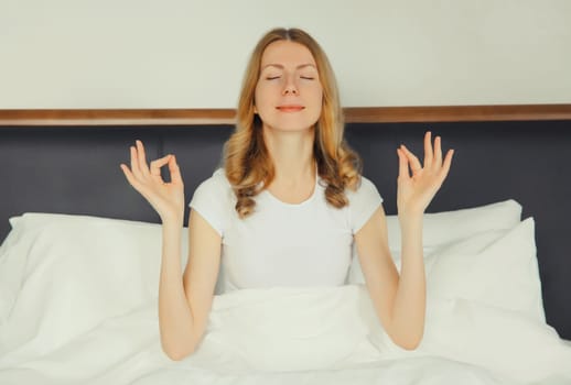 Calm relaxed healthy young woman meditates, girl practicing doing yoga exercises sitting in lotus pose on white bed at home, stress relief