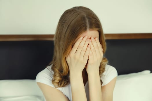 Upset sad crying young woman covering her eyes and face with her hands while experiencing depression, stress, fatigue, mental suffering after sleeping sitting on the bed at home